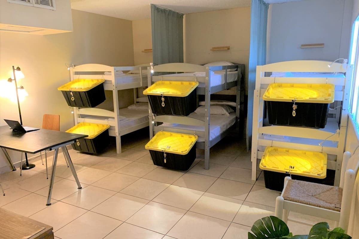 A dormitory room with bunk beds and storage bins, a desk with a laptop, and a chair.