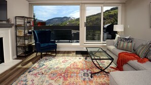 Un salon avec une cheminée, un fauteuil bleu, une table basse en verre, un canapé gris et un tapis coloré.