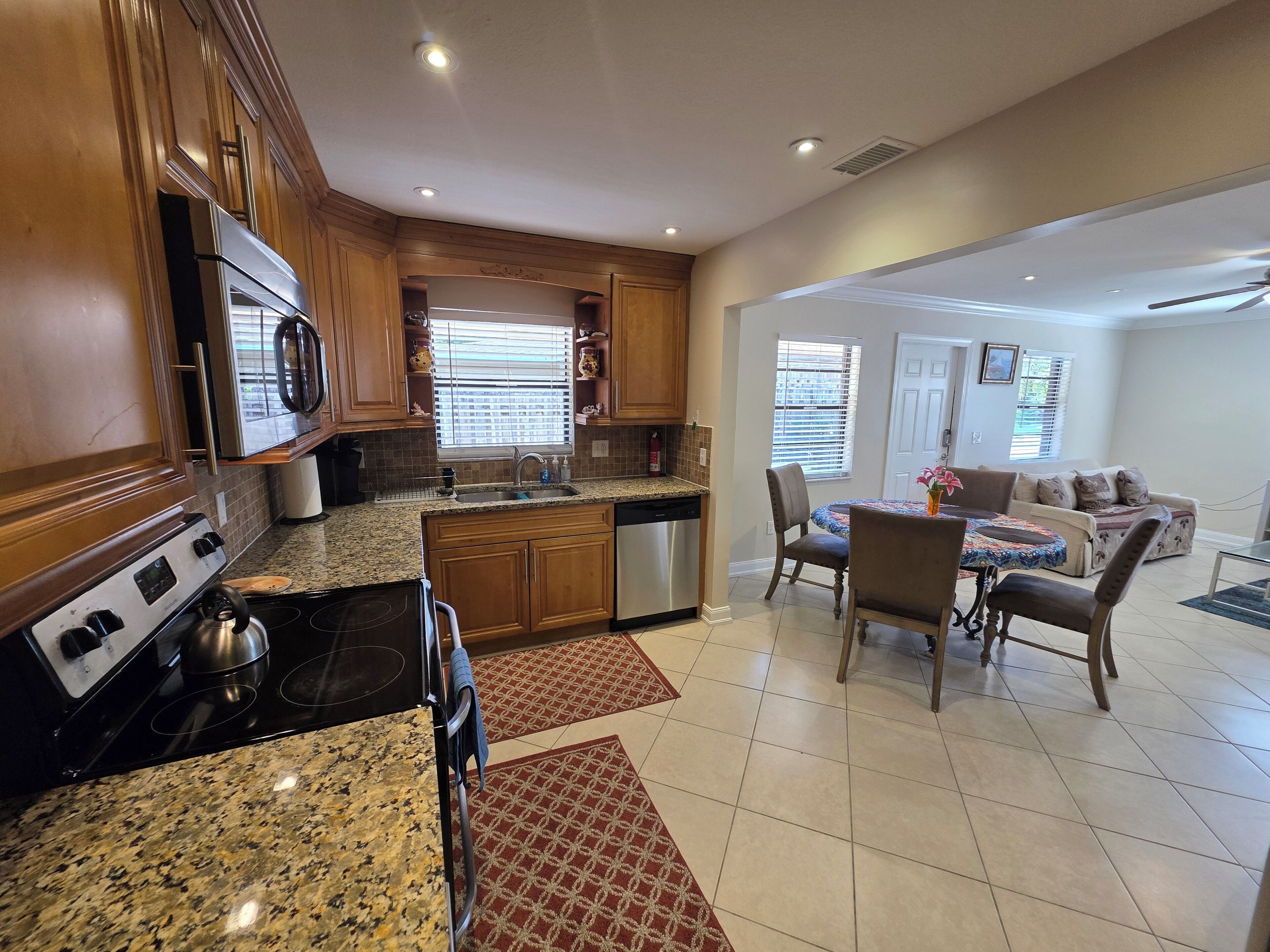 A modern kitchen with granite countertops, wooden cabinets, and a central island. There is a dining area with a table and chairs, and a living room with a sofa and a TV.