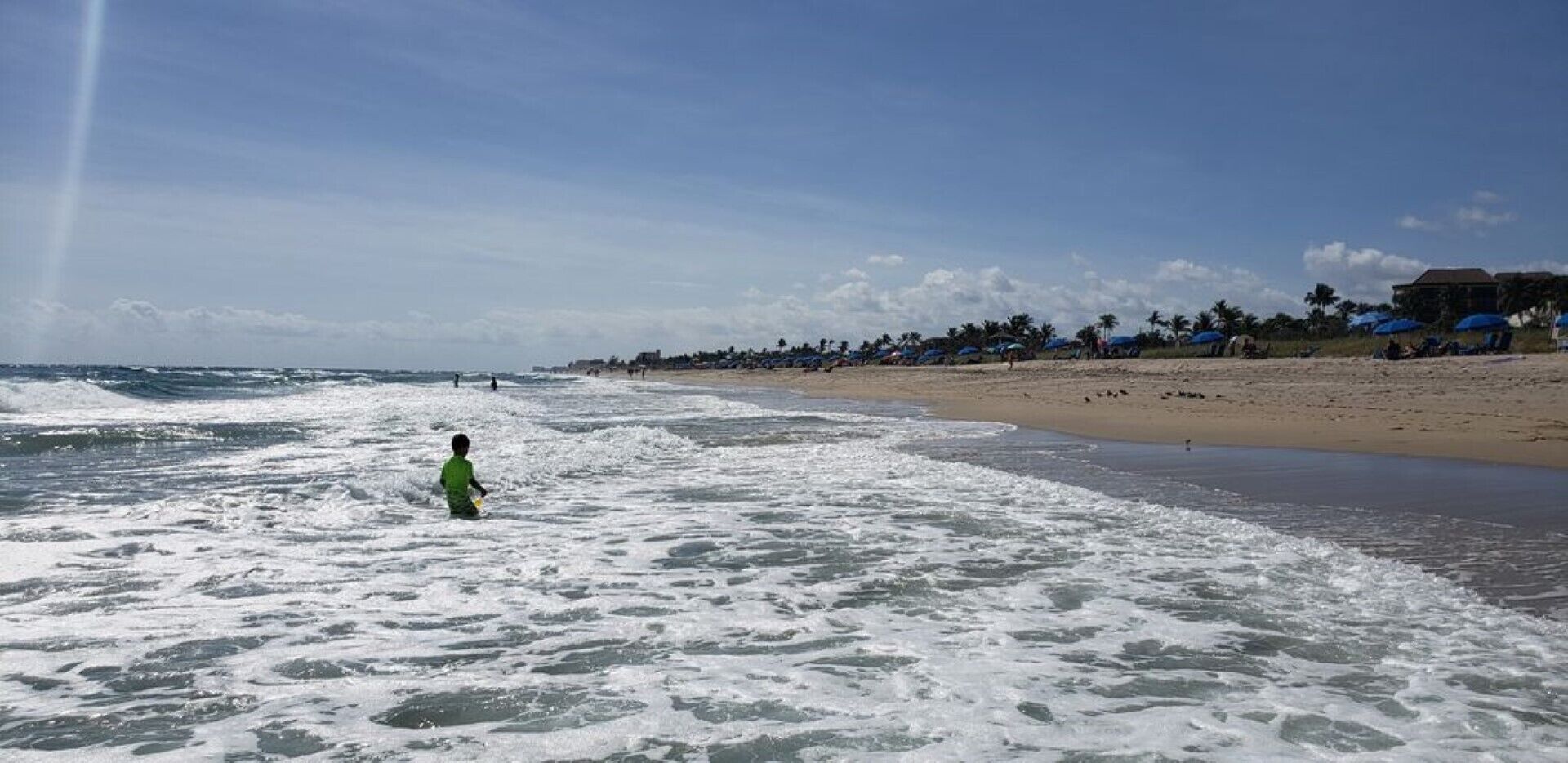 Una playa con olas, un niño en el agua y sombrillas de playa al fondo.