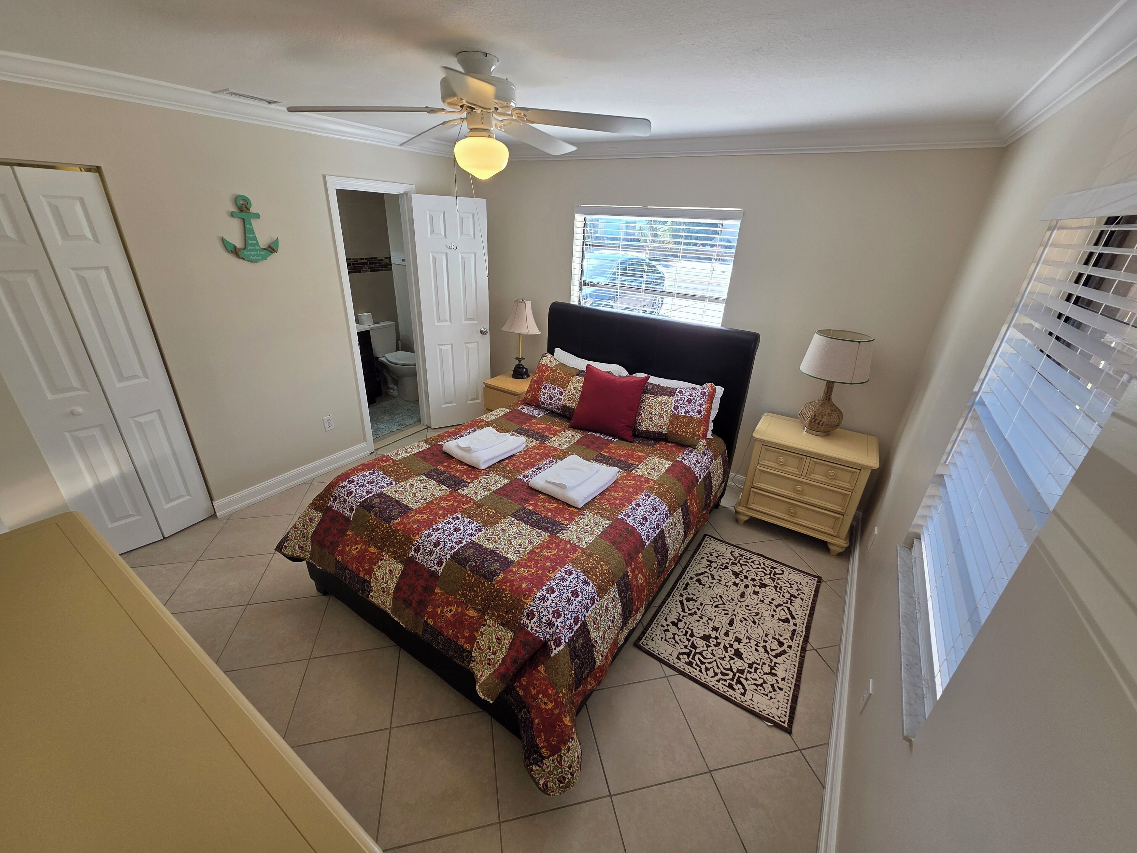 A bedroom with a bed, a ceiling fan, a window with blinds, a nightstand, and a door leading to a bathroom.