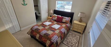 A bedroom with a bed, a ceiling fan, a window with blinds, a nightstand, and a door leading to a bathroom.