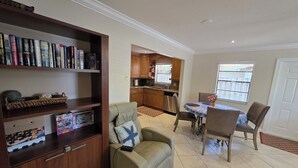 Een woonkamer met een bank, een salontafel en een boekenkast. In de achtergrond bevindt zich een keuken met houten kastjes en een raam.