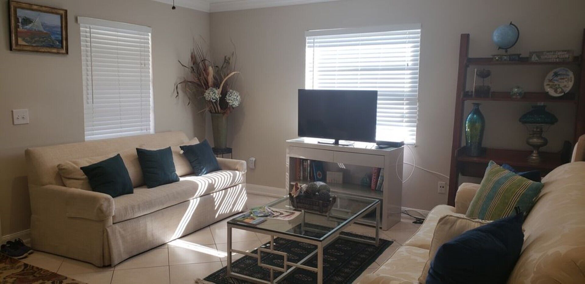Ein Wohnzimmer mit einem beigefarbenen Sofa, einem Glastisch, einem Fernseher auf einem Schrank und einem Bücherregal mit Dekoration.