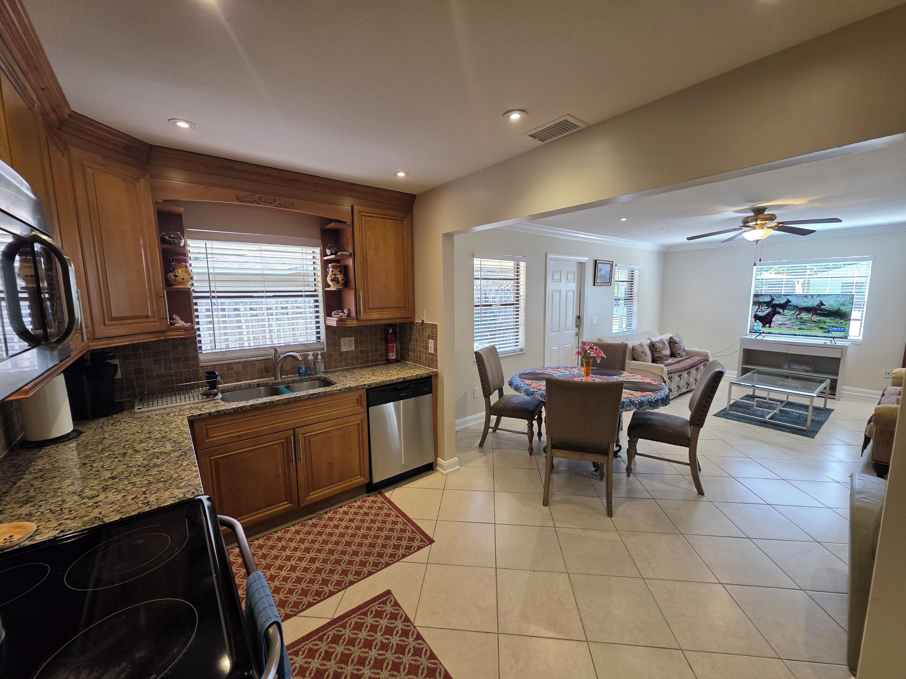 A modern kitchen with granite countertops, wooden cabinets, and stainless steel appliances. There is a dining area with a round table and chairs, and a living room with a sofa and a television.
