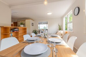 A dining area with a wooden table set for a meal, surrounded by white chairs. There is a bookshelf with books and a potted plant on the left. - Redwoods Haven - Spa Pool, Sleeps 9 (Rotorua)