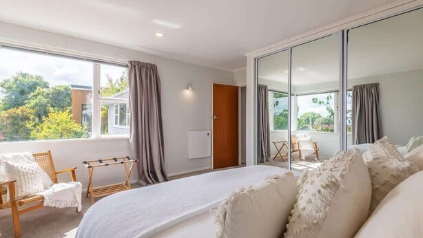 A bedroom with a large bed, a view of the outdoors, a wooden door, and a window with curtains. - Redwoods Haven - Spa Pool, Sleeps 9 (Rotorua)