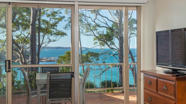 A room with a large sliding glass door leading to a balcony with a view of the sea and trees. - Windarra on Denhams Unit Two (DENHAMS BEACH)