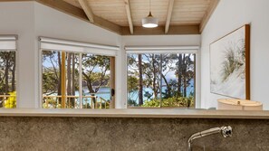 A modern bathroom with large windows, a wooden ceiling, and a view of trees and water.