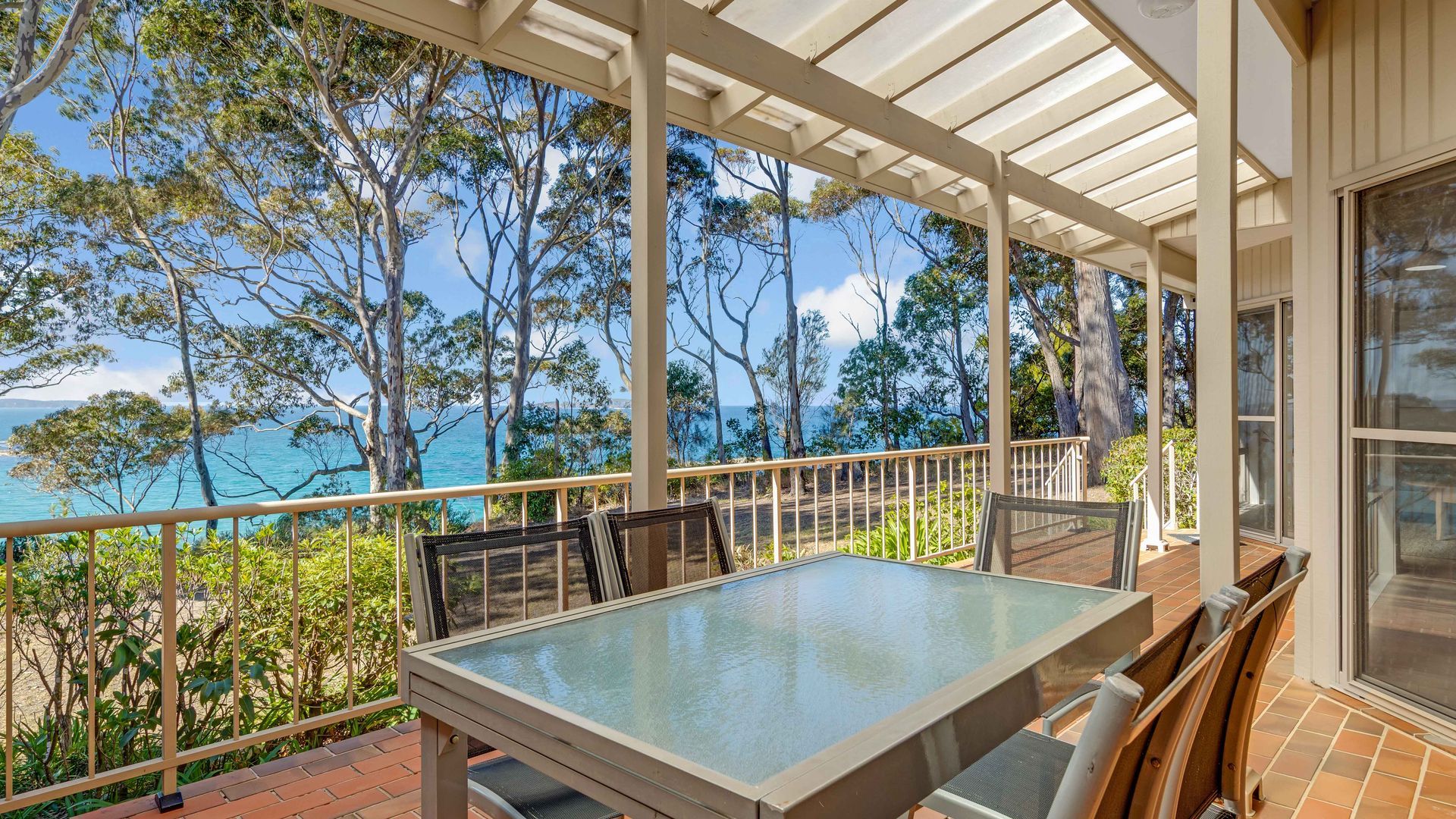 A covered outdoor area with a table and chairs, overlooking a coastal landscape with trees and the sea.