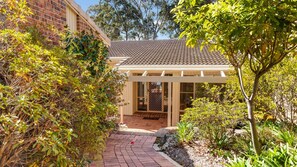A covered outdoor area with a tiled roof, surrounded by lush greenery and a brick pathway leading to a building entrance. - Windarra on Denhams Unit Two (DENHAMS BEACH)