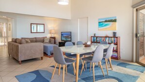 A living room with a sofa, a TV, a dining table with chairs, a bookshelf, and a painting of a beach. - Windarra on Denhams Unit Two (DENHAMS BEACH)