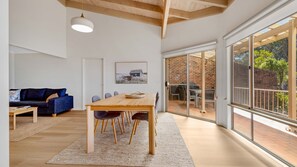 A spacious room with a wooden dining table, chairs, and a large window offering a view of the outdoors. - Windarra on Denhams Unit Two (DENHAMS BEACH)
