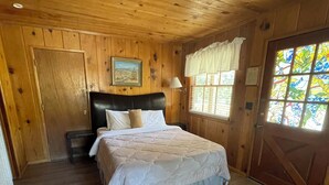 Un dormitorio de cabaña de madera con cama, mesita de noche, ventana con cortinas y puerta con diseño de vitral.