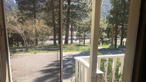 Vista a través de una ventana de un porche con barandilla de madera, que da a un área pavimentada con árboles y una cerca.