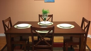 Uma área de refeições com uma mesa e cadeiras de madeira, um tapete com padrão vermelho e branco, e duas pinturas emolduradas de paisagens na parede.