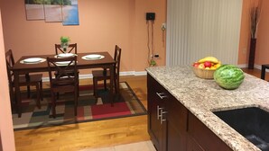 Uma cozinha com bancada de granito, armários de madeira, um espaço de jantar com mesa e cadeiras, e uma tigela de frutas no balcão.