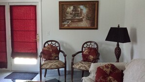 A room with two wooden chairs with patterned cushions, a white sofa, a framed painting on the wall, a lamp, and a red door. - Cozy Apartment on Cape Cod MA (Mashpee)