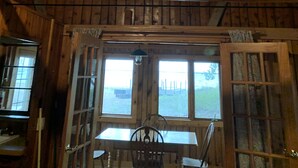 A rustic wooden interior with a dining table and chairs, a ceiling fan, and large windows offering a view of the outdoors. - (Birch Cabin) Waterfront 2 Bed 1 Bath Cottage - Pets OK (East Tawas)
