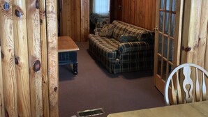 A wooden cabin interior with a plaid sofa, a dining table, and a window with curtains. - (Birch Cabin) Waterfront 2 Bed 1 Bath Cottage - Pets OK (East Tawas)