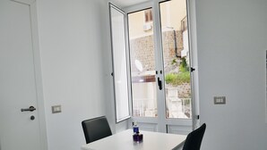 Une petite pièce avec une table blanche et deux chaises, une fenêtre donnant sur un bâtiment en pierre, et une porte avec une poignée.