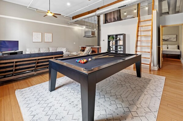 Une table de billard dans un salon moderne, avec un canapé, un téléviseur et un escalier menant à une mezzanine.