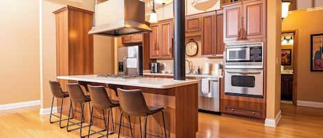 Una cocina con armarios de madera, una isla central con taburetes de bar, electrodomésticos de acero inoxidable e iluminación colgante.