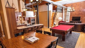 A dining area with a wooden table, chairs, and a red pool table. There is a kitchen area with cabinets and a stove in the background.