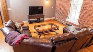 A living room with leather sofas, a glass coffee table, a flat-screen TV, and a brick wall.