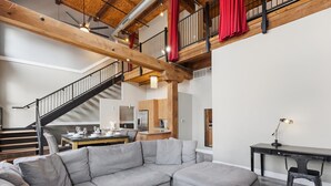 Ein modernes Wohnzimmer mit einer Eckcouch, einem Essbereich mit Tisch und Stühlen, einer Küche im Hintergrund und einer Treppe mit roten Vorhängen.