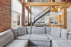 A modern loft apartment with exposed brick walls, a large gray sectional sofa, a kitchen area, and a staircase leading to an upper level.