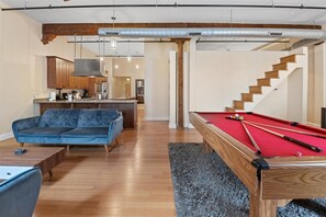 A living room with a pool table, a blue sofa, wooden flooring, and a staircase leading to a kitchen area. - Top Floor Loft in Downtown Indianapolis!!! (Indianapolis)