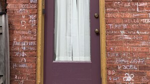 Eine violette Tür mit weißem Vorhang, ein kleines Fenster darüber und eine Ziegelwand mit Graffiti.