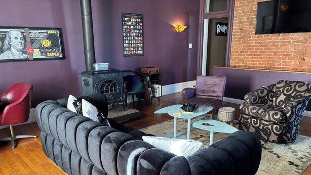Ein Wohnzimmer mit einer schwarzen Couch, einem roten Sessel, einem gemusterten Sessel, einem kleinen Tisch, einem Kamin und einem Poster an der Wand.