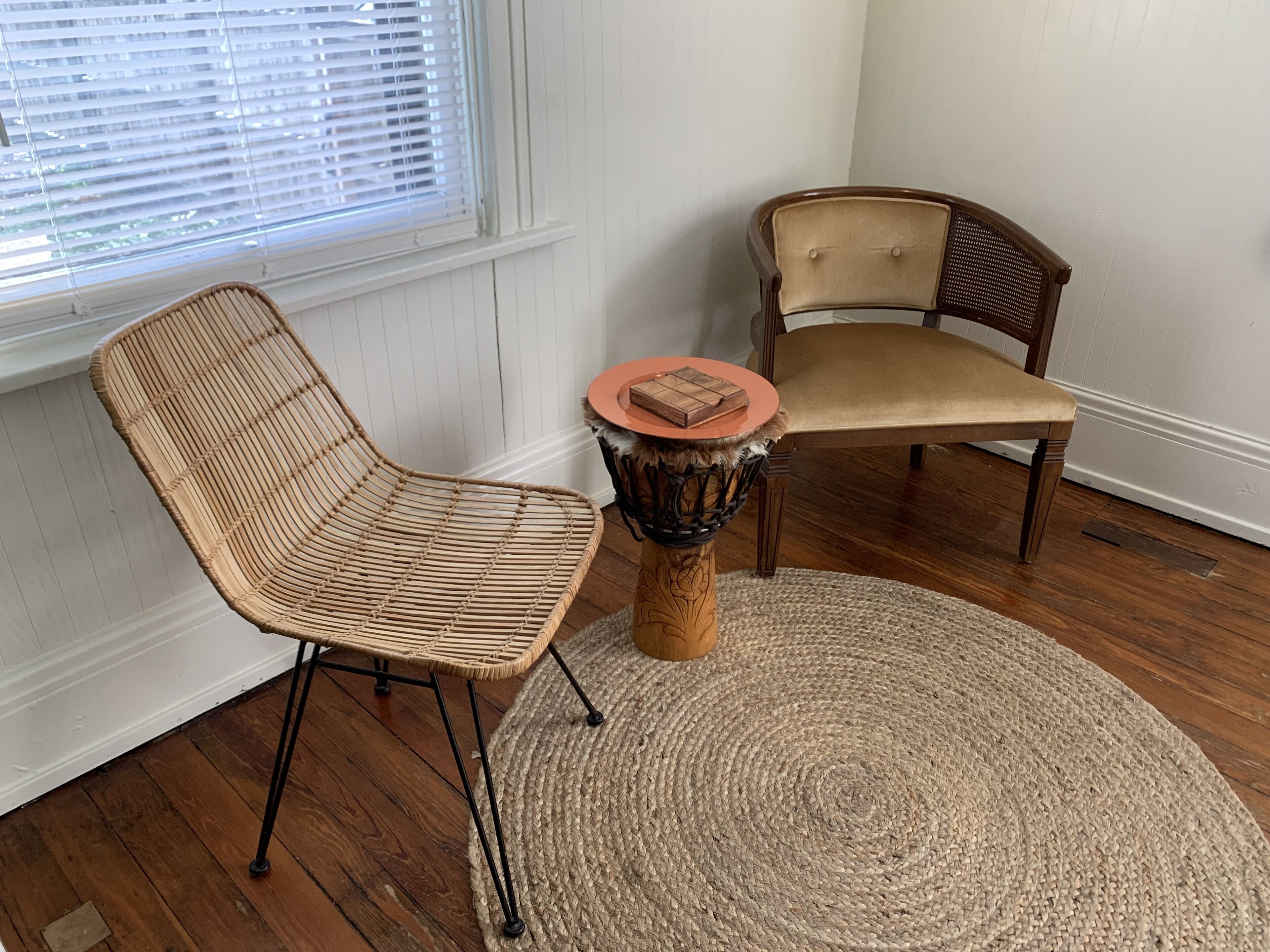 Een kamer met een rieten stoel, een houten stoel en een kleine ronde tafel met een geweven vloerkleed.