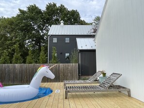 Un terrazzo in legno con poltrone relax e una piscina, un unicorno gonfiabile bianco e un edificio scuro sullo sfondo.