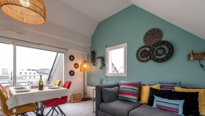 Uma sala de estar moderna com um sofá cinzento, almofadas coloridas, uma mesa de jantar com cadeiras, um abajur e uma janela grande.