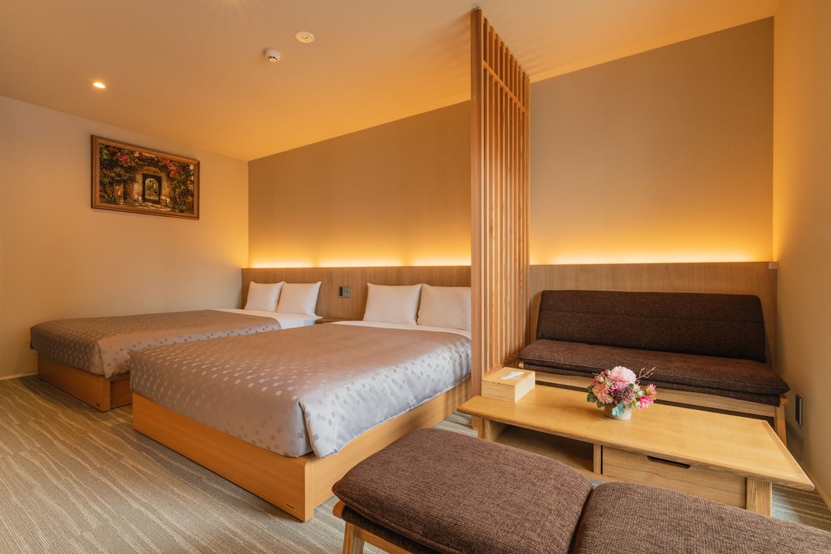 Ein Hotelzimmer mit zwei Betten, einem Sofa, einem kleinen Tisch mit Blumenvase und einem Bild an der Wand.