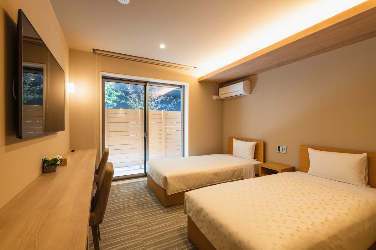 Ein Hotelzimmer mit zwei Einzelbetten, einem Schreibtisch mit Stuhl, einem an der Wand montierten Fernseher und einer Schiebetür zum Balkon.