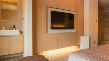 A modern hotel room with a large flat-screen TV mounted on the wall, a wooden vanity with a mirror, and a small white appliance, likely a mini-fridge, on the floor.