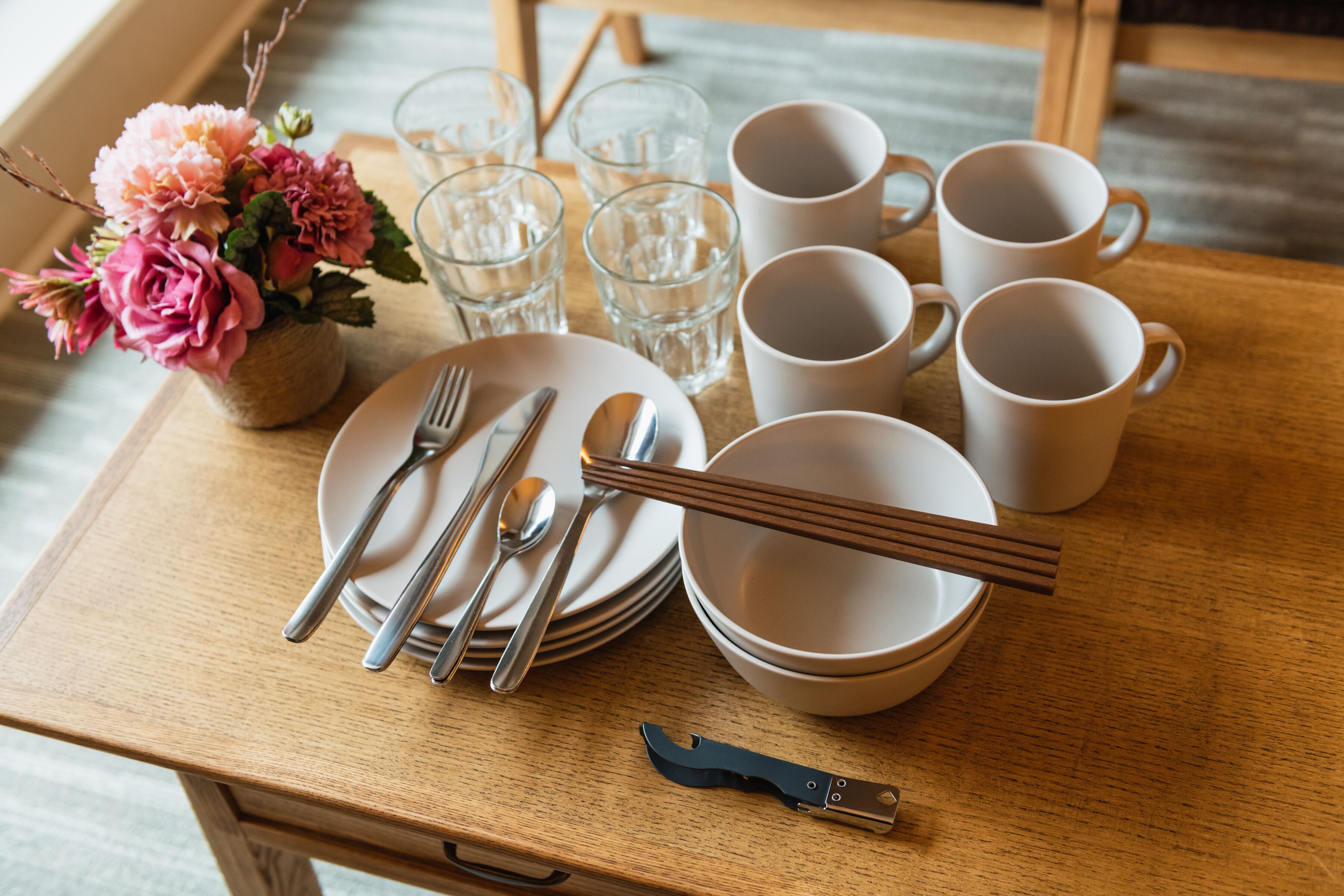 Ein Holztisch mit weißen Tellern, Schüsseln und Besteck, begleitet von rosa Blumen und klaren Gläsern.