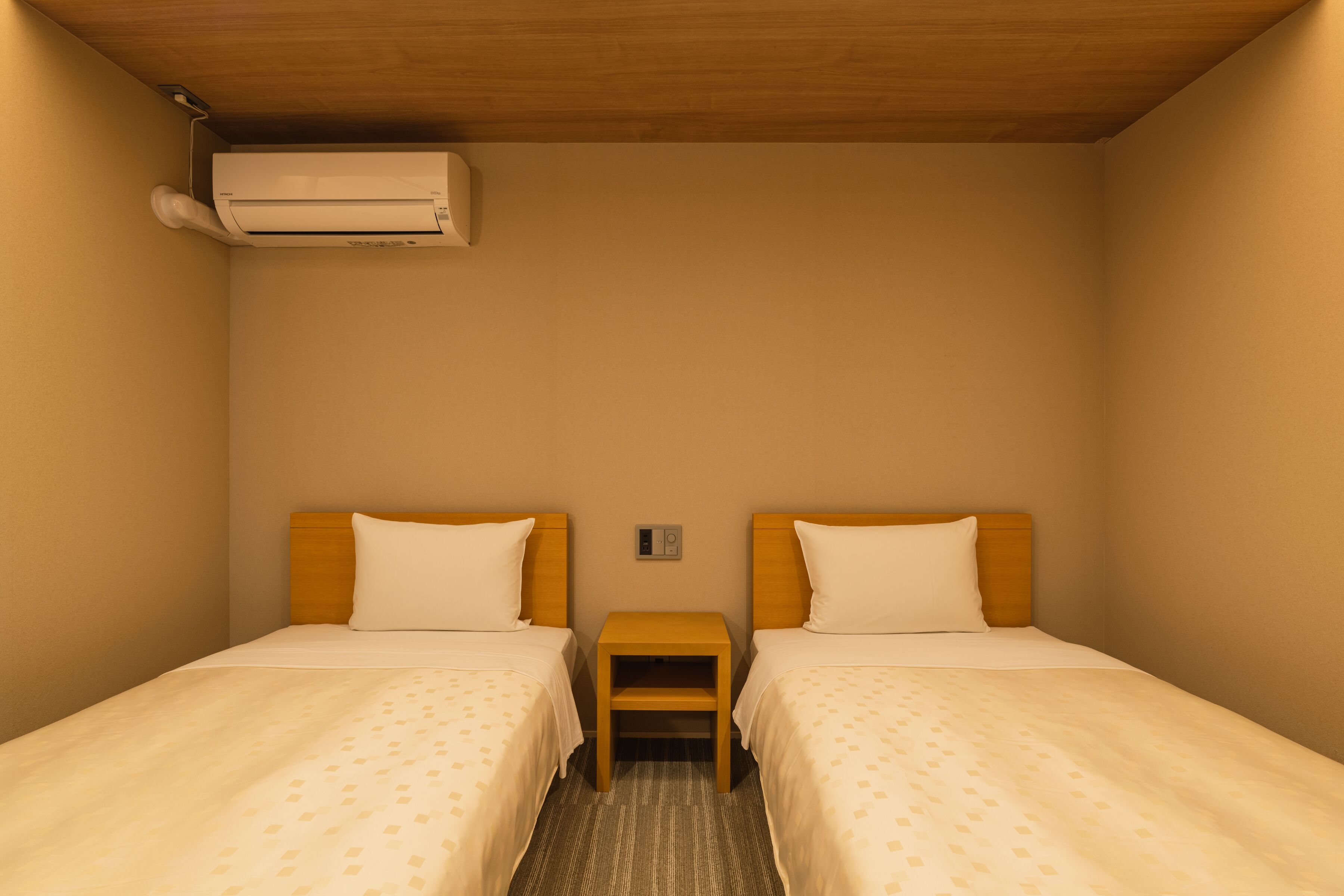 A hotel room with two beds, a wooden headboard, a small nightstand, and a wall-mounted air conditioner.