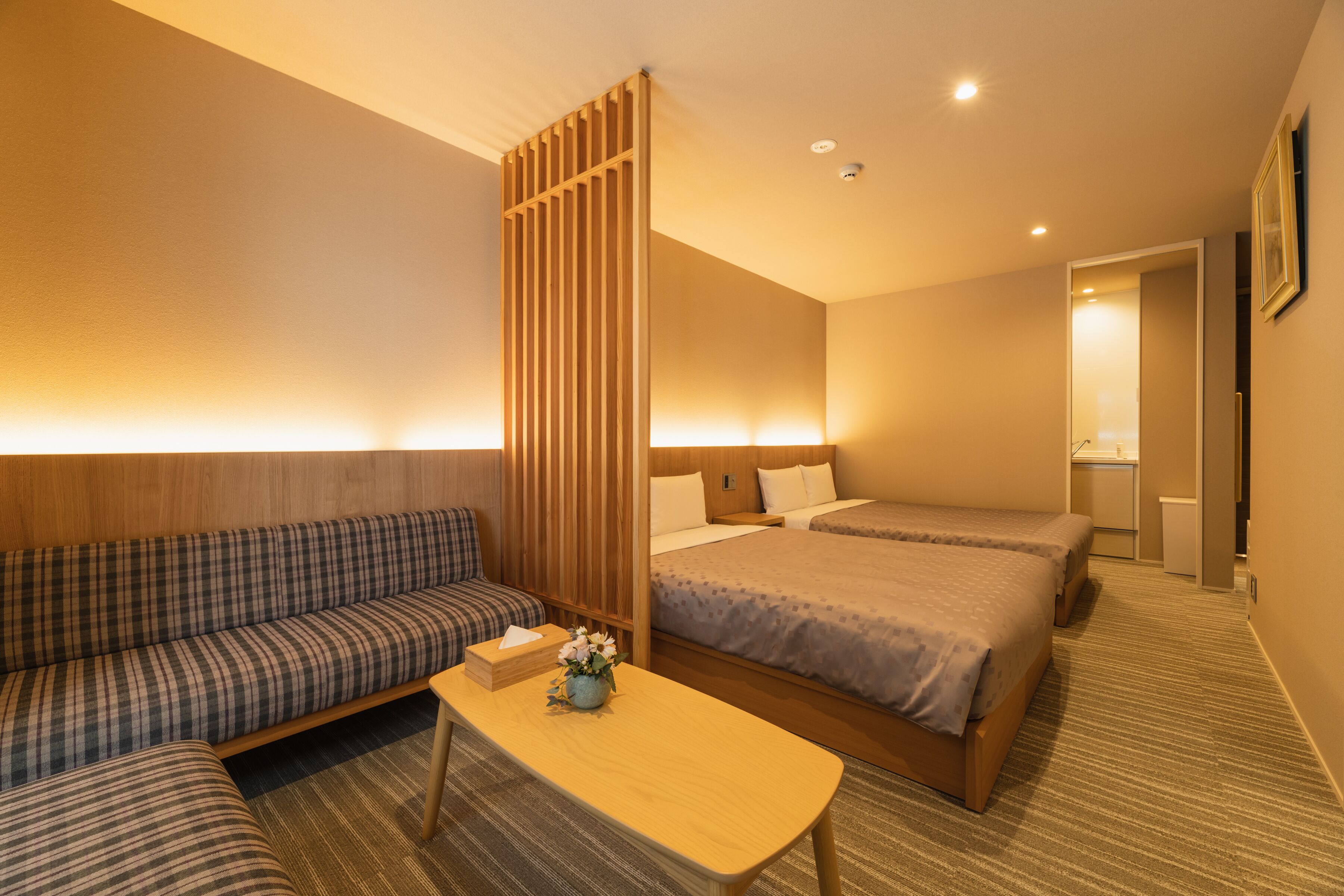 A hotel room with a sofa, two beds, a wooden partition, and a small table with a vase.