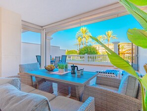 Une terrasse extérieure avec des meubles en osier, une table avec une cafetière et une vue sur des palmiers et des immeubles.