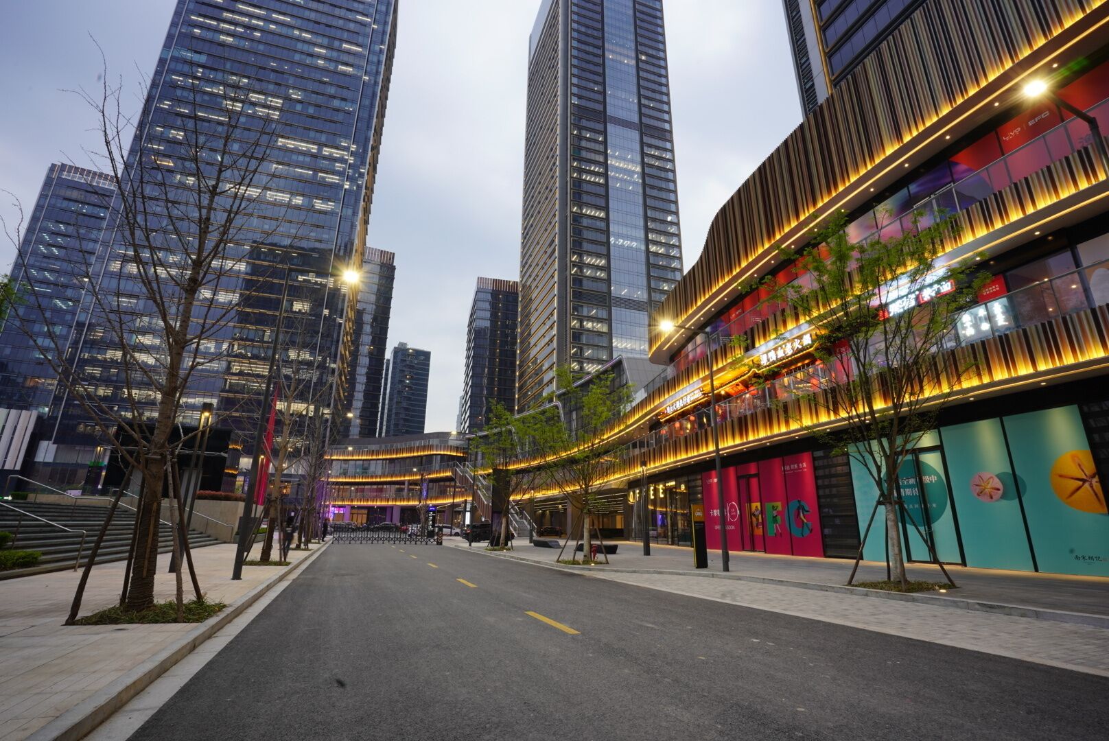 A modern urban street with high-rise buildings, a pedestrian walkway, and a brightly lit commercial building with signage.