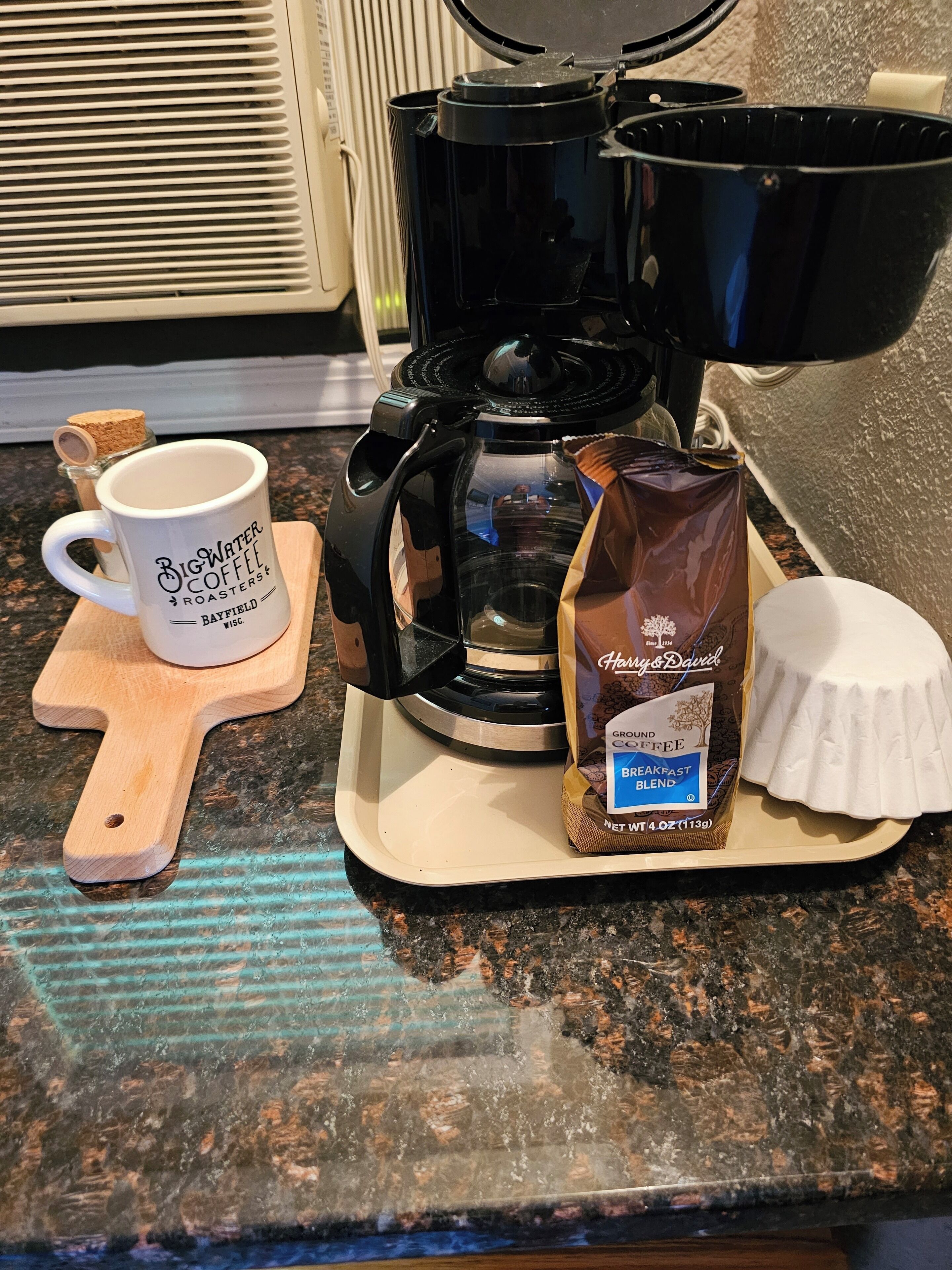 A coffee maker, a coffee mug, a packet of ground coffee, and a scoop of creamer on a kitchen counter.