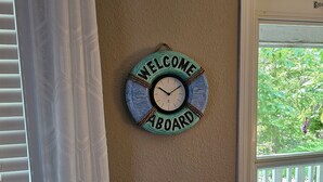 A wall clock with "WELCOME ABOARD" on a textured wall near a window with blinds.