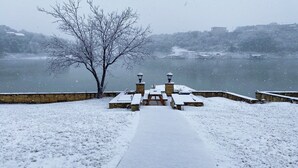 Área externa com neve, bancos e uma parede de pedra, com vista para um corpo de água.