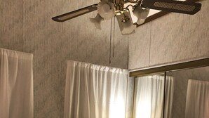 Quarto de hotel com uma cama grande, um ventilador de teto, mesa de cabeceira com abajur e espelho de corpo inteiro.