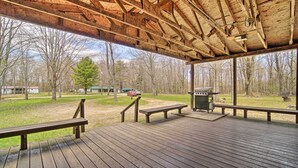 A covered wooden deck with benches and a grill area, overlooking a grassy area with trees and a distant building.